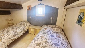 Quarto pequeno e compacto com duas camas de solteiro, uma cadeira de madeira, uma mesinha e uma janela com vista.