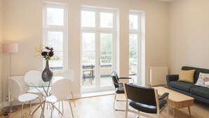 A modern living room with a glass dining table, white chairs, a black leather sofa, and a view through large windows. - Beautiful 2 bed terrace flat close to Greenwich -  Pass the Keys (London)