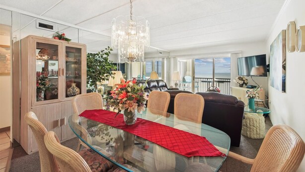 Un comedor amplio con una mesa de vidrio, sillas de mimbre y un corredor rojo. Un gran candelabro cuelga encima, y la habitación tiene vista al mar a través de amplios ventanales.