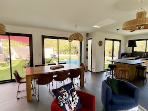 Un vaste espace de vie comprenant un coin repas avec une table, des chaises rouges et bleues, un coin cuisine et une vue sur un jardin avec une piscine.