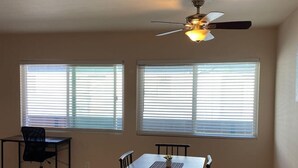 Un comedor con mesa y sillas, ventilador de techo y ventanas amplias con persianas.