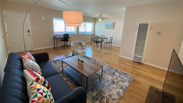 Una sala de estar con un sofá de color oscuro, una mesa de centro de madera y un tapete estampado. Hay una zona de comedor con una mesa y sillas, un ventilador de techo y una ventana con persianas.