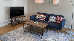 Una sala de estar con televisor sobre un mueble bajo, un sofá con cojines estampados, una mesa de centro y una lámpara de pie.