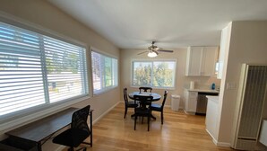 A spacious room with a dining table and chairs, a desk, a ceiling fan, and large windows with blinds.