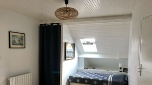 Ein kleines Schlafzimmer unter dem Dach mit freundlichem Licht, geneigter Decke, einem Bett mit Sternenmuster, einem kleinen Regal mit Dekoration und einem Bilderrahmen an der Wand.