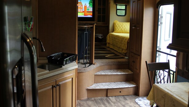 A compact kitchen with a refrigerator, sink, and a small countertop. There is a window with a view of the outside, and a bed with a yellow blanket in the adjacent sleeping area. - Luxury 5th Wheel Glamping In Style In Historic Mentone,  Alabama (Mentone)