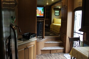 A compact kitchen with a refrigerator, sink, and a small countertop. There is a window with a view of the outside, and a bed with a yellow blanket in the adjacent sleeping area. - Luxury 5th Wheel Glamping In Style In Historic Mentone, Alabama (Mentone)