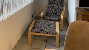 Um quarto com uma janela coberta por cortinas leves, uma cadeira com almofada estampada, uma mesa pequena e um quadro na parede.