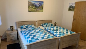 Uma cama de casal com roupa de cama quadriculada azul e branca, uma cabeceira de madeira, uma mesa de cabeceira com um abajur, uma pequena gaveta e um tapete preto.