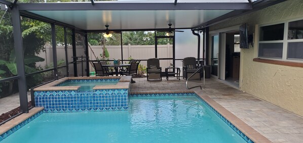 A covered outdoor swimming pool with a patio area, including a table and chairs, and a view of the backyard.