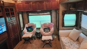 A cozy RV interior with wooden cabinets, two cushioned chairs with pillows, a small table, and a sofa with a floral bedspread.