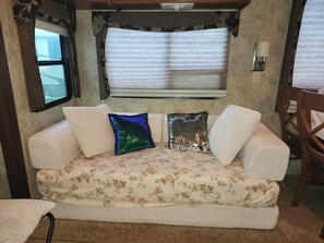 A cozy interior with a sofa, decorative pillows, a window with blinds, and a wall-mounted lamp. - Luxury 5th Wheel Glamping In Style In Historic Mentone,  Alabama (Mentone)