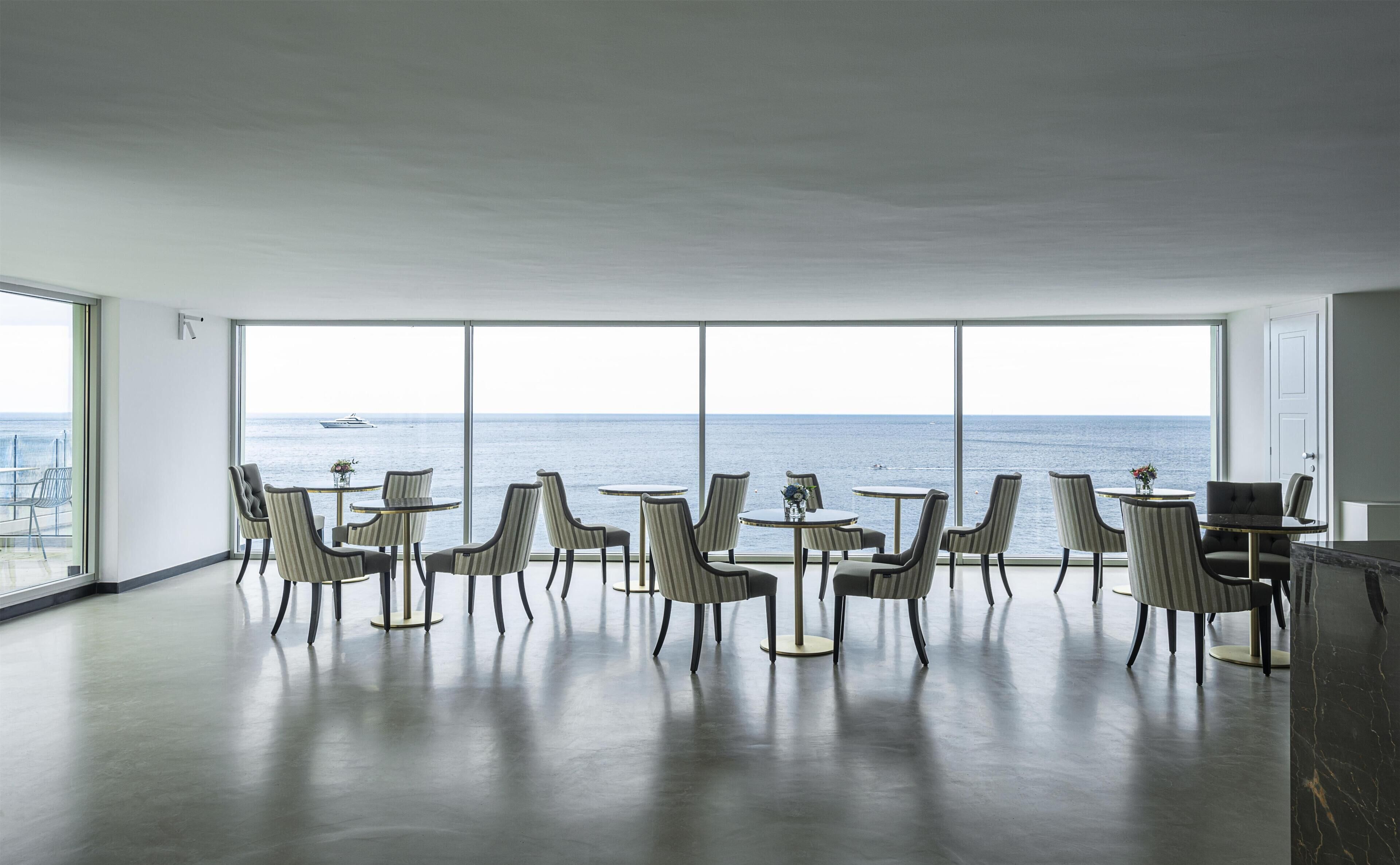 A spacious room with large windows, a view of the sea, and a dining area with chairs and tables.