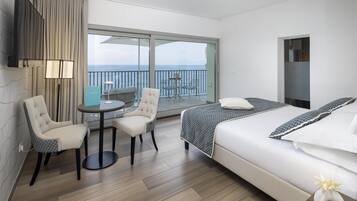 A hotel room with a bed, a small table, two chairs, a TV, and a balcony with a view of the sea.