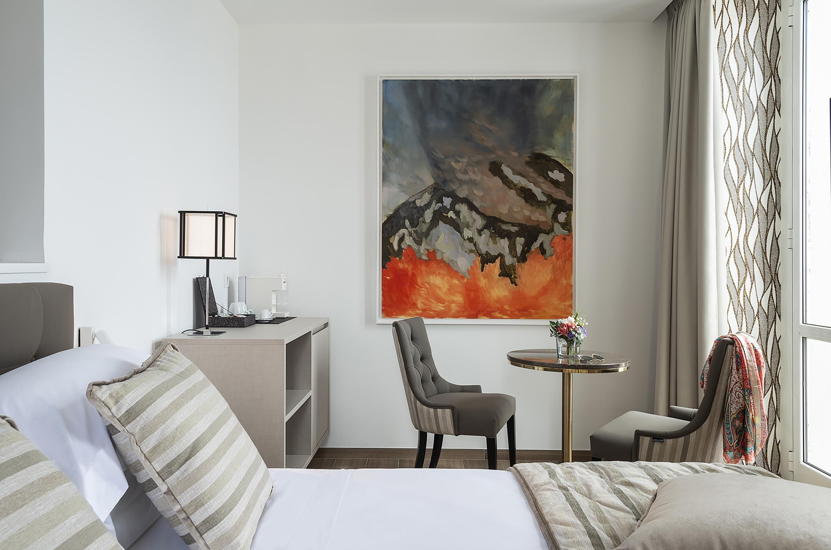 A hotel room with a bed, two chairs, a small table, a desk, and a large abstract painting on the wall.