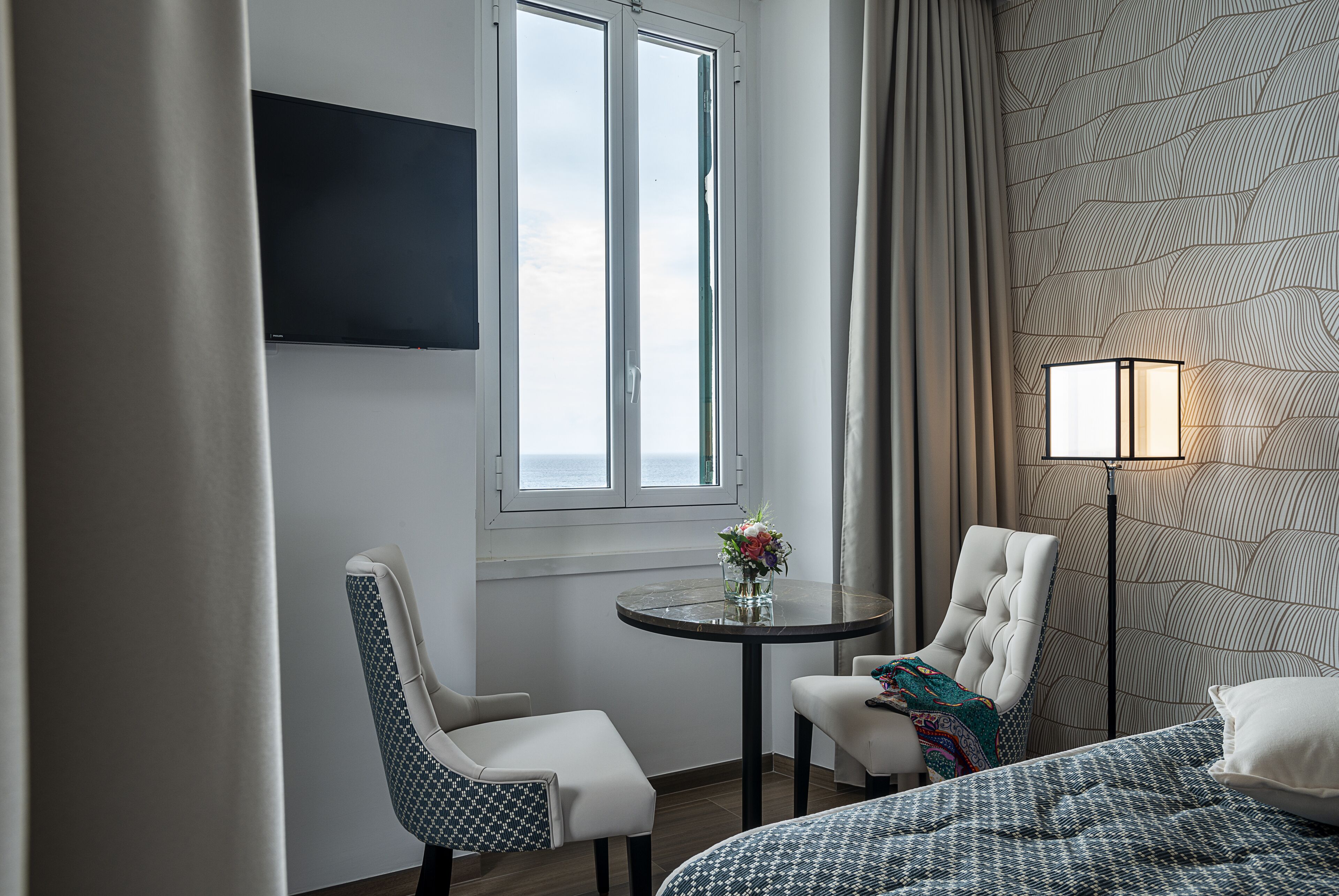 A hotel room with a bed, two chairs, a small table with a vase of flowers, a TV mounted on the wall, and a patterned wallpaper.