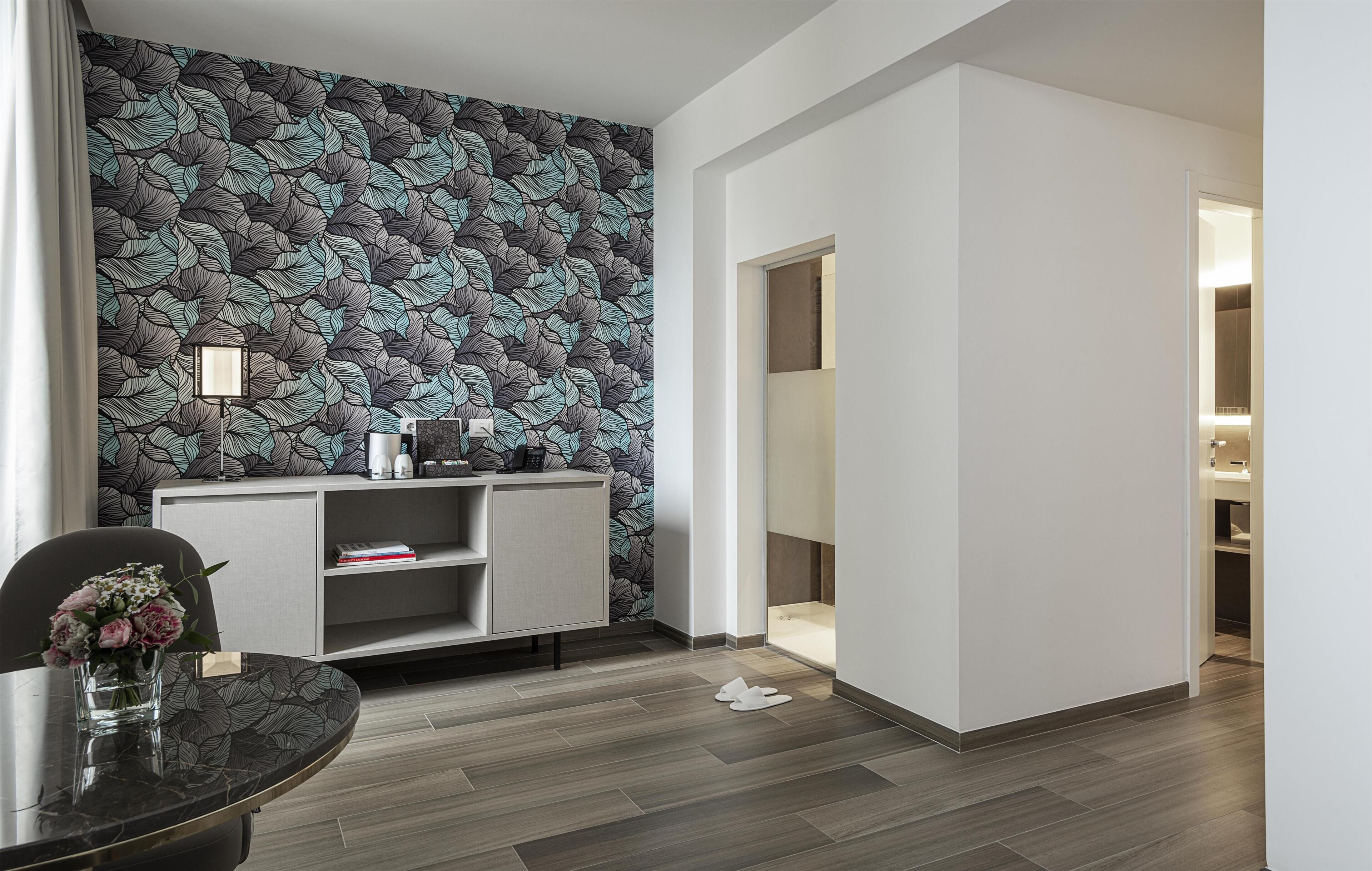 Ein modernes Hotelzimmer mit gemustertem Wandteppich, einem grauen Konsolentisch, einem runden Esstisch mit Blumenvase und einem sichtbaren Badezimmer.