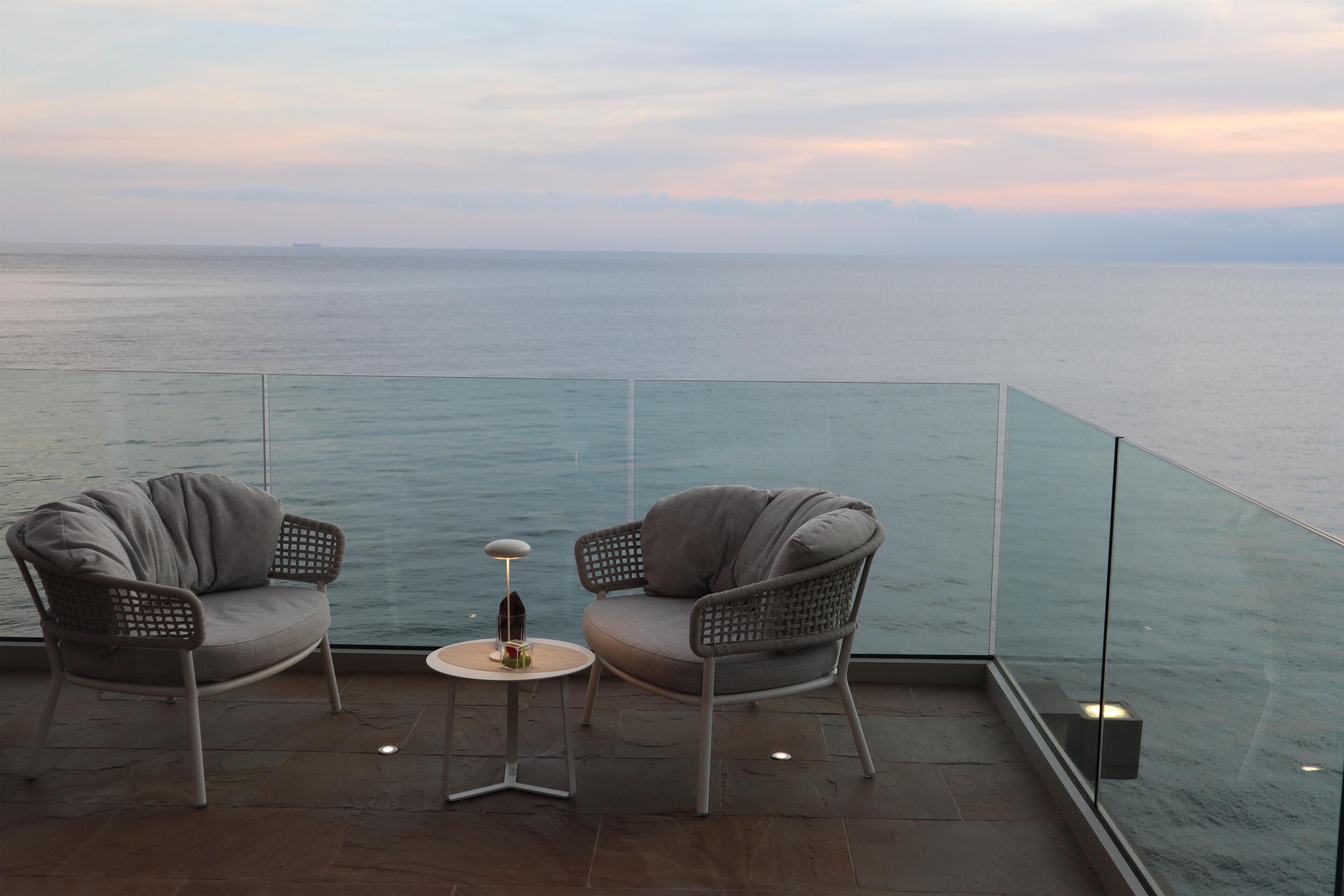 A balcony with two grey chairs and a small table, overlooking a calm sea.