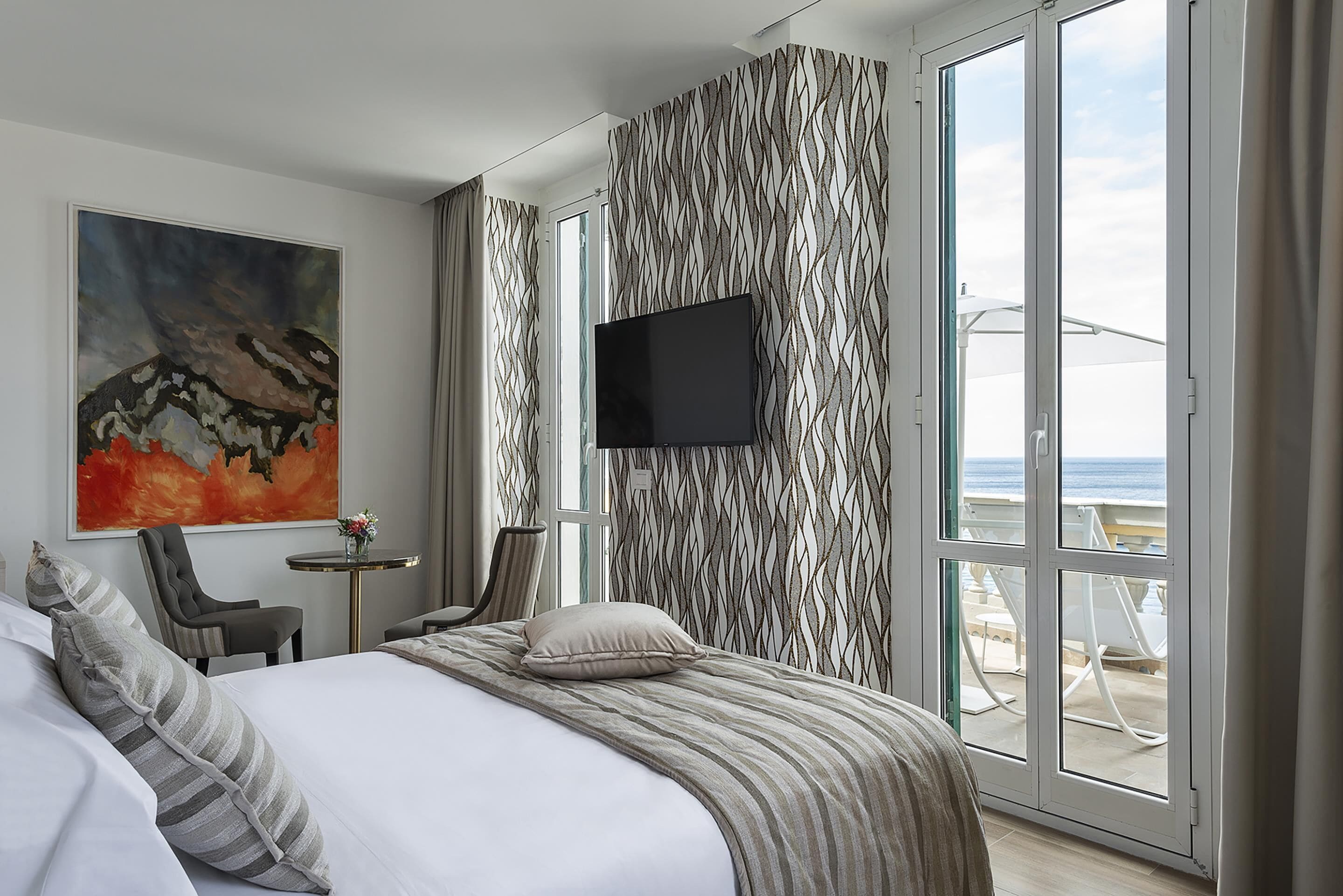 Une chambre d’hôtel équipée d’un lit, d’une télévision, d’une chaise, d’une petite table et offrant une vue sur la mer grâce à de grandes fenêtres.