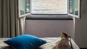 Een kamer met een bed, een raam met uitzicht op zee en een tas op het bed.