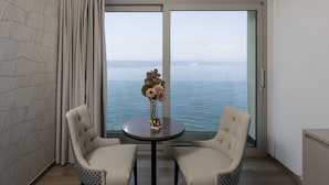 Een kamer met een groot raam met uitzicht op zee, twee stoelen, een klein tafeltje met een vaas en een kamer met een patroon op de muur.
