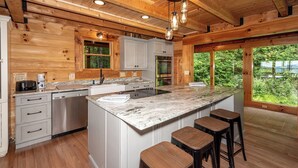 A modern kitchen with wooden walls, a large island with bar stools, stainless steel appliances, and a view of greenery. - This 5 bedroom 5 bath log cabin with spectacular views, secluded, & Hot-tub (Townsend)