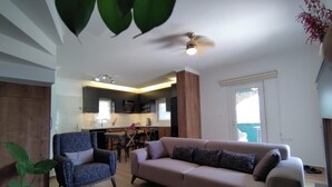 A living room with a sofa, armchair, and a coffee table with magazines. A kitchen is visible in the background with a dining area.