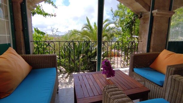 A covered outdoor seating area with wicker furniture and cushions, a wooden table, and a view of greenery through an archway.