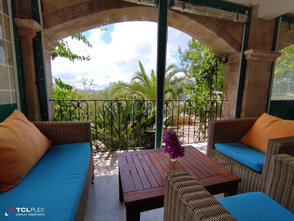 Un espace extérieur aménagé, abrité et doté de mobilier en osier et de coussins, d’une table en bois et d’une vue sur la verdure, visible à travers une arche.