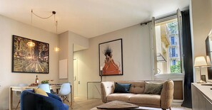 A modern living room with a sofa, a coffee table, and a dining area with a chair and a table. There are two paintings on the wall, and a window with a view of buildings.