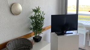 En moderne stue med et fladskærms-tv på et hvidt skab, en potte med bambus, dekorativ vægkunst og et stort vindue med udsigt udendørs.