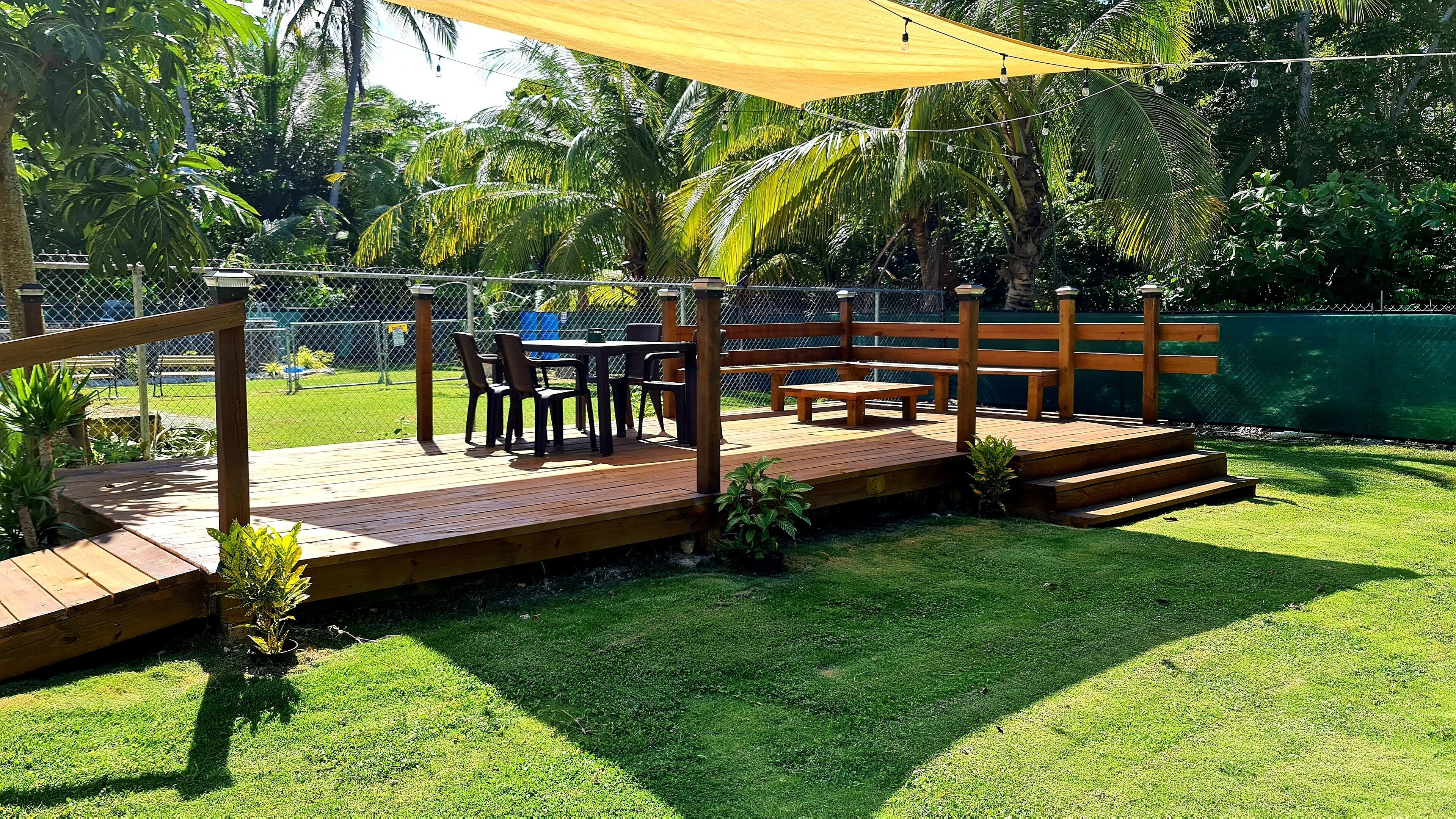 Una terraza de madera con una mesa y sillas, rodeada por una cerca de malla y abundante vegetación.