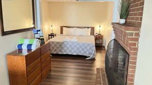 Una habitación de hotel con una cama, tocador de madera, mesitas de noche, chimenea y espejo.
