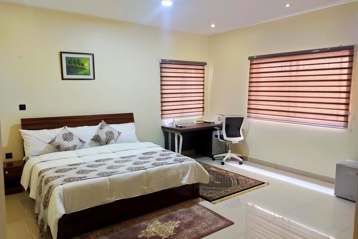 A bedroom with a bed, desk, chair, and two windows with blinds.