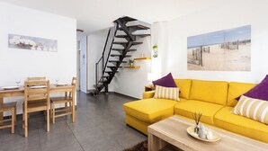 Una sala de estar moderna con un sofá amarillo, una mesa de comedor de madera con sillas, una escalera espiral negra y un cuadro con motivos de playa en la pared.