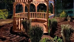 Um gazebo de madeira com telhado curvo e grade, rodeado por canteiros ajardinados e árvores.