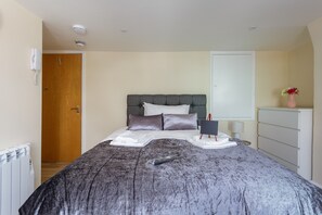 Una cama bien hecha con edredón gris, una mesita de noche con lámpara y una puerta de madera al fondo.