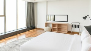 A modern bedroom with a large bed, a wooden floor, a grey rug, a window with curtains, a wooden console table, a black lamp, and a wall-mounted radiator. - Dara Hotel Qurtubah (Riyadh)