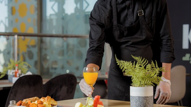 A person serving food in a modern dining area with a decorative plant and a glass of orange juice on the table. - Dara Hotel Qurtubah (Riyadh)