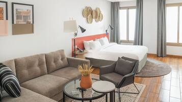 A modern hotel room with a bed, sofa, armchair, and a small coffee table.
