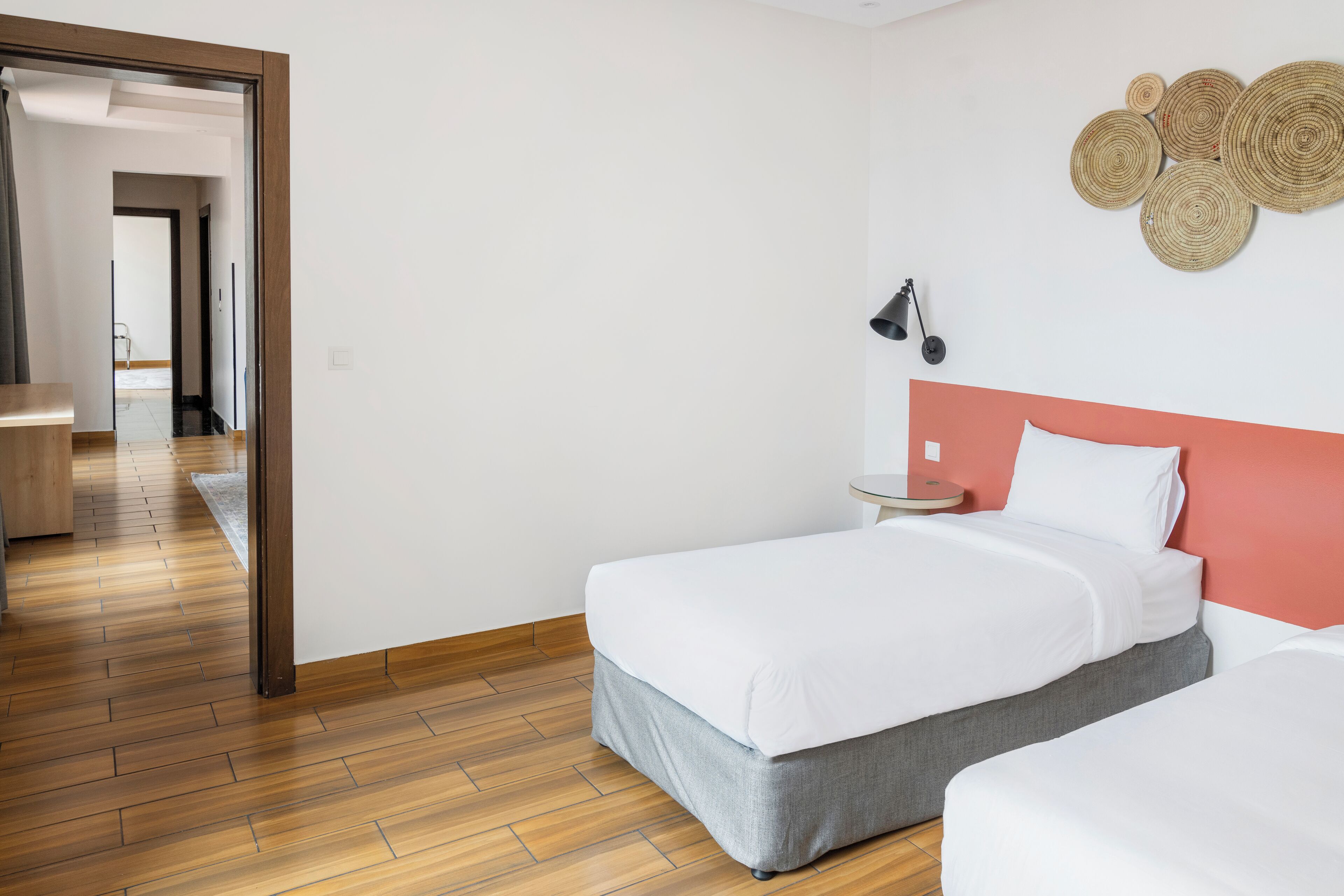 A hotel room with a wooden floor, a bed with a grey skirt, a white headboard, and a wall-mounted lamp.