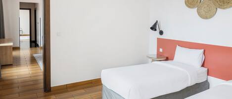 A hotel room with a wooden floor, a bed with a grey skirt, a white headboard, and a wall-mounted lamp.