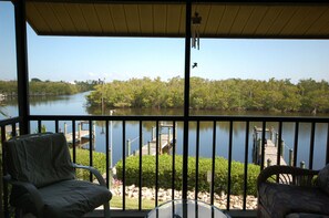 A balcony with two chairs, a table, and a view of a calm river with a dock and trees. - Captain's Harbor Condo Relax: on your private screened balcony and enjoy the scenery just outside this gorgeous condo. (Bokeelia)