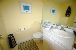 A bathroom with a toilet, sink, and mirror. There are framed pictures on the wall and a small shelf with toiletries. - Corner Condo in Blue Crab Key, Bokeelia! (Saint James City)