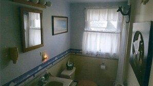 A small bathroom with a sink, toilet, and mirror. There is a window with a curtain and a picture on the wall. - Quiet Getaway Within Walking Distance of the Beach and Mall. (Cape May)