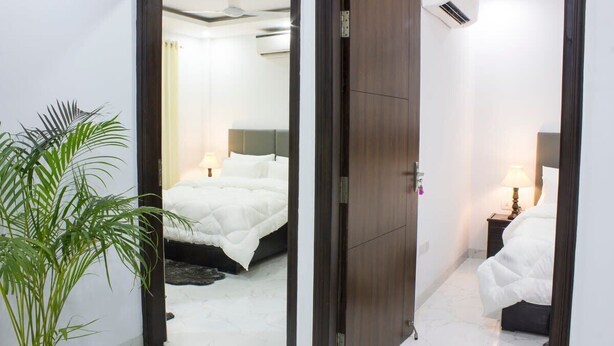 A hotel room with a bed, bedside table, lamp, and a potted plant. - Central Stay, Medical Ease 2BHK (Gurugram)