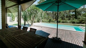 Eine Holzterrasse mit Tisch und Stühlen, einem grünen Sonnenschirm und einem Pool im Hintergrund.