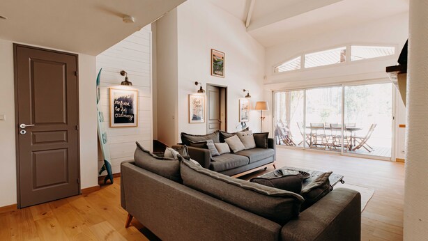 Ein modernes Wohnzimmer mit einem grauen Ecksofa, Holzboden und einer großen Glasschiebetür, die auf eine Terrasse im Freien führt.