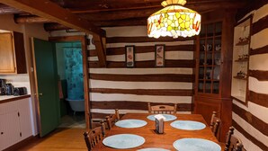 Una mesa de comedor de madera con cuatro individuales, rodeada de sillas de madera, en una habitación con vigas de madera a la vista y una lámpara con detalles de vidrio coloreado.