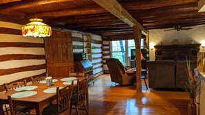 Interior rústico de una cabaña con una mesa de madera, sillas, una araña, una chimenea y una zona de cocina.